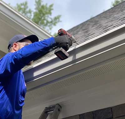 Person installing a gutter on a house roof with power drill.