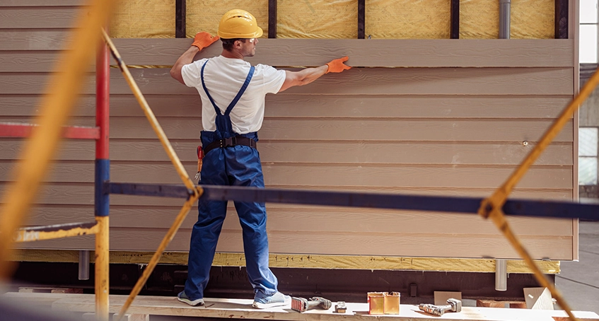 Worker in overalls and helmet installing house siding panels.