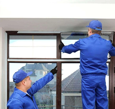 Two workers in blue uniforms installing a large window panel indoors.